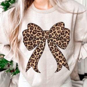 May include: A beige sweatshirt featuring a large bow design with a leopard print pattern in shades of brown and tan. The bow is centered on the front of the shirt, creating a bold and stylish look. The sweatshirt has a crew neck and long sleeves.