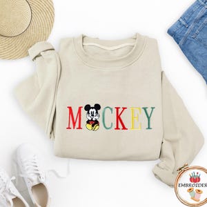 May include: A beige sweatshirt with a red, green, and yellow embroidered Mickey Mouse design and the word 'MICKEY'.