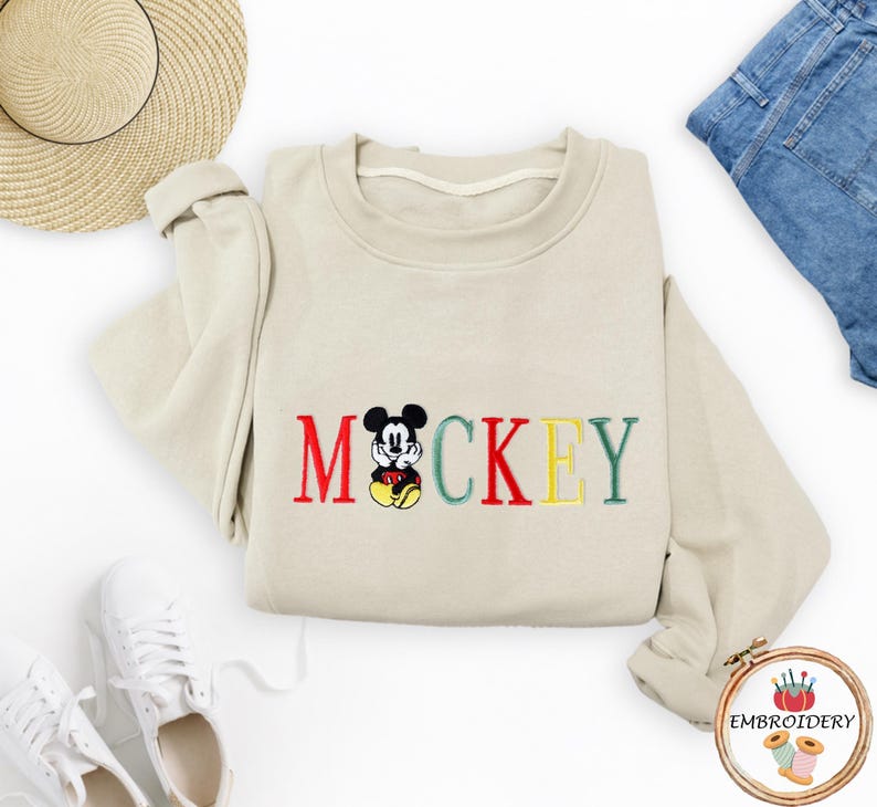 May include: A beige sweatshirt with a red, green, and yellow embroidered Mickey Mouse design and the word 'MICKEY'.