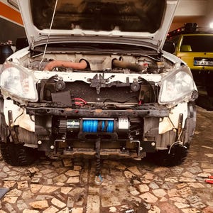 May include: A white pickup truck with the hood open, revealing the engine. The truck has a black winch mounted on the front bumper. The winch has a blue cable and a green and black label. The truck is parked in a garage with a yellow SUV behind it.