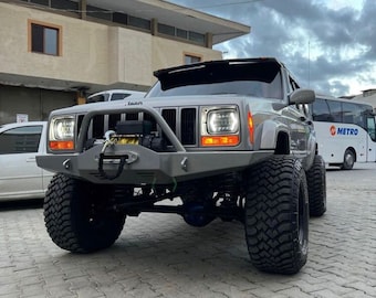 May include: A gray Jeep Cherokee with a black front bumper and a winch. The Jeep has large tires and is parked on a brick driveway. The Jeep is in front of a building with a minaret.