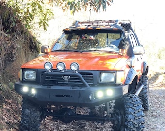 May include: An orange Toyota pickup truck with a black roof rack and a black front bumper. The truck has large off-road tires and is parked on a dirt road.