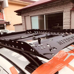 May include: A black metal roof rack with the word "MITSUBISHI" embossed on the side. The rack is fitted to a silver car.