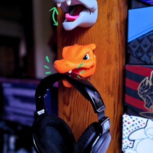 May include: A pair of black over-ear headphones hanging on a wooden surface. Two small plastic figurines of cartoon characters are attached to the surface above the headphones. The figurine on the top is blue and the figurine on the bottom is orange.