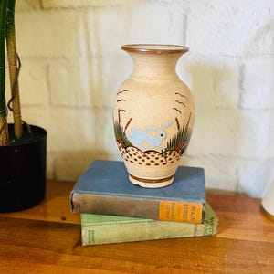 May include: A beige ceramic vase with a hand-painted design of a blue rabbit, brown grass, and a brown rim. The vase is sitting on top of two vintage books. A green plant and a desk lamp are in the background.