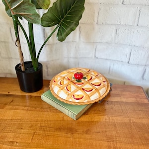 Könnte beinhalten: Eine Keramik-Tortenform mit Deckel, verziert mit einem roten Apfel und einer Gitterkruste. Die Form ist mit gelben und roten Apfelscheiben gefüllt. Die Form steht auf einem grünen Buch auf einem Holztisch.