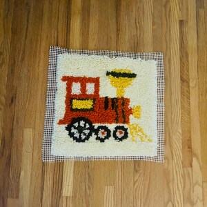 Puede incluir: Un proyecto de manualidades de ganchillo con cierre cuadrado que presenta un diseño de tren sobre una base de malla blanca. El tren es principalmente naranja con una chimenea amarilla, ruedas negras con centros blancos y vapor amarillo.