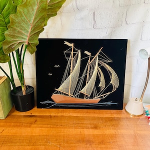 May include: A framed artwork featuring a sailboat crafted with gold thread on a black background. The boat has tan sails and a brown hull. Two small birds are depicted in the sky. The artwork is displayed on a wooden surface.