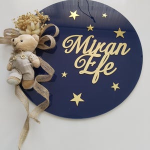 May include: A navy blue circular wall decoration with gold stars and the name "Miran Efe" in gold script. A small teddy bear and dried flowers are attached with a burlap ribbon. A string is attached for hanging.