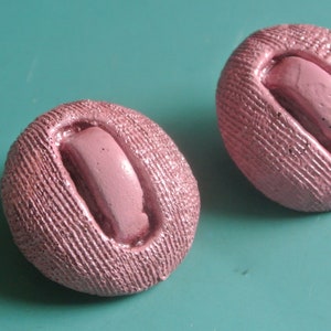 May include: Two pink fabric covered buttons with a raised oval design in the center. The buttons have a textured surface.
