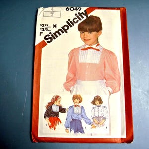Op de afbeelding: Een Simplicity 6049 naaipatroon-envelop met een perzikkleurige blouse met lange mouwen, een wit geplooid voorpand en een rode strik. De omslag toont illustraties van vier variaties van de blouse in verschillende kleuren en patronen.