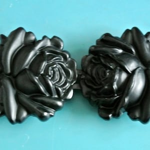 Puede incluir: Un par de hebillas de cinturón de plástico negro en forma de rosa. Las hebillas están detalladas con múltiples capas de pétalos.