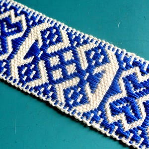 Könnte beinhalten: Nahaufnahme eines gewebten Textils mit einem sich wiederholenden geometrischen Muster in Blau und Weiß. Das Design weist stilisierte Formen und ein strukturiertes Aussehen auf, wahrscheinlich für dekorative Zwecke oder als Besatz verwendet.