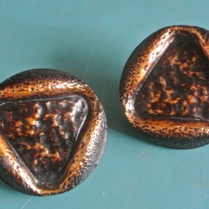 May include: A pair of round, copper-toned earrings with a textured, triangular design. The earrings have a dark brown finish with a copper-colored border.