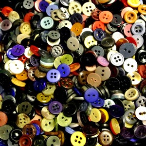 May include: A large collection of assorted sewing buttons in various colors, shapes, and sizes. The pile includes round buttons with two or four holes, featuring shades of black, white, blue, red, yellow, green, brown, and grey.
