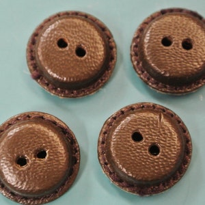 Puede incluir: Cuatro botones redondos marrones con una superficie texturizada y dos agujeros cada uno. Los botones tienen un borde cosido marrón oscuro. Los botones están dispuestos sobre una superficie azul claro.