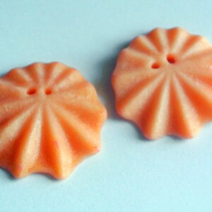 May include: Two orange, flower-shaped buttons with radial grooves and two holes each. The buttons have a slightly textured surface and are set against a light blue background. These buttons are ideal for sewing and craft projects.