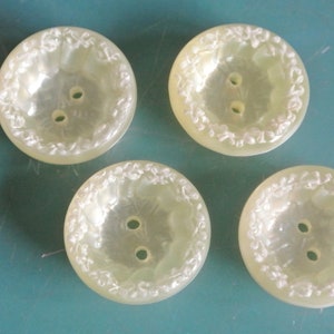 Puede incluir: Cuatro botones de plástico de color blanco roto con un diseño floral texturizado en relieve alrededor del borde. Los botones tienen dos agujeros cada uno.