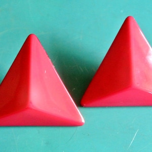 Pode incluir: Dois objetos vermelhos brilhantes em forma de pirâmide. São lisos e brilhantes, com uma base triangular e três lados que se encontram num ponto. Colocados sobre um fundo turquesa.