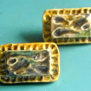 May include: Two rectangular ceramic cabinet knobs. Each knob has a textured, yellow-brown border and a central rectangle with a mottled design in shades of green, gray, and brown. The knobs are set against a teal background.