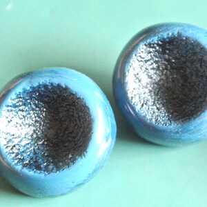 May include: Two round, button-like objects featuring a smooth, glossy, bright blue outer rim and a deep, concave center with a dark, textured, matte black finish. The objects have a spherical shape with a distinct indentation in the middle.