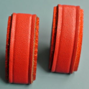 May include: Two red leather-covered bookends with a decorative border. The bookends are rectangular in shape and have a smooth, textured surface.