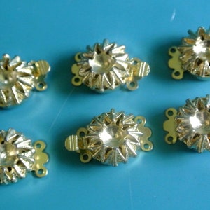 Puede incluir: Seis cierres de metal dorado con un diseño decorativo en forma de estrella. Cada cierre tiene dos bucles para sujetar a joyas u otros artículos.