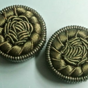 Könnte beinhalten: Zwei dunkelgrüne, stoffbezogene Knöpfe mit einem silbernen Metallrand. Die Knöpfe haben ein gewebtes Design, das an eine Blume erinnert.