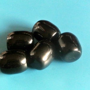 Könnte beinhalten: Eine Ansammlung von polierten, schwarzen Steinen mit glänzender Oberfläche. Die Steine sind oval geformt und liegen auf einem hellblauen Hintergrund. Es handelt sich um eine Sammlung von Edelsteinen oder Dekorationsobjekten.