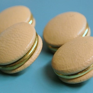 May include: Four beige macarons with a gold band around the middle. The macarons are arranged in a semi-circle on a blue surface.