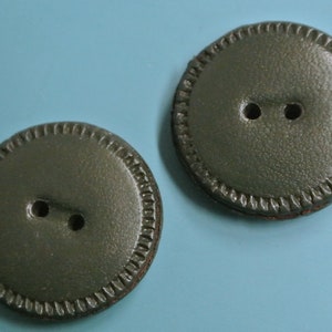 Puede incluir: Dos botones de cuero verde oscuro con un borde cosido decorativo. Cada botón tiene dos agujeros.