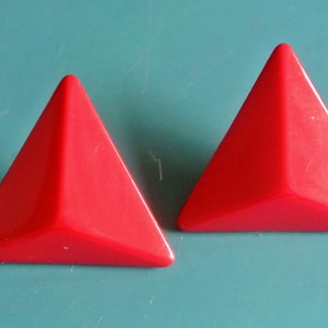 May include: A pair of red triangular shaped earrings. The earrings are made of a shiny plastic material.