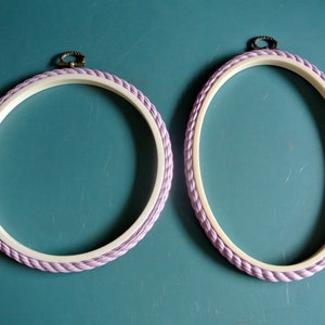 May include: Two embroidery hoops with a light purple rope border and a white inner ring. The hoops are on a teal surface.
