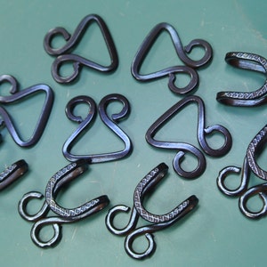 Op de afbeelding: Tien zwarte metalen haak- en oogsluitingen. De haken hebben een decoratief patroon en de ogen zijn driehoekig.