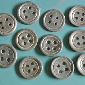Peut inclure: Un ensemble de onze boutons circulaires aux tons métalliques, chacun présentant un centre en retrait et quatre trous régulièrement espacés pour la fixation. Les boutons ont une finition mate et patinée, disposés de manière éparse.
