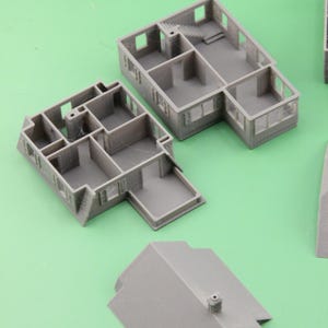 Op de afbeelding: Grijze 3D-geprinte miniatuurhuismodellen, inclusief muren, vloeren en een dak. De modellen hebben gedetailleerde kenmerken zoals ramen, deuren en trappen. De stukken zijn gerangschikt op een groene ondergrond, wat het architectonisch ontwerp laat zien.