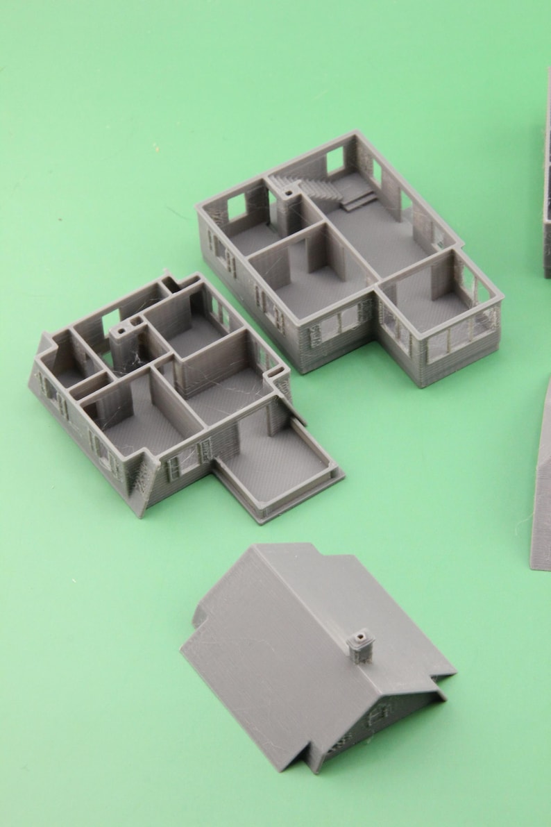 Op de afbeelding: Grijze 3D-geprinte miniatuurhuismodellen, inclusief muren, vloeren en een dak. De modellen hebben gedetailleerde kenmerken zoals ramen, deuren en trappen. De stukken zijn gerangschikt op een groene ondergrond, wat het architectonisch ontwerp laat zien.