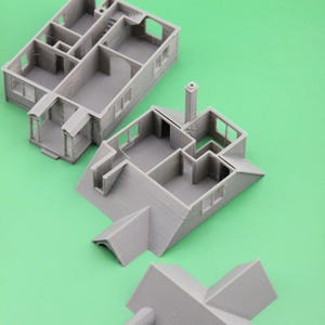 Op de afbeelding: Grijs 3D-geprint modelhuis in verschillende gedemonteerde secties, inclusief muren, dak en schoorsteen. Het model toont interieurdetails zoals kamers en trappen. De stukken zijn gerangschikt op een groene ondergrond.