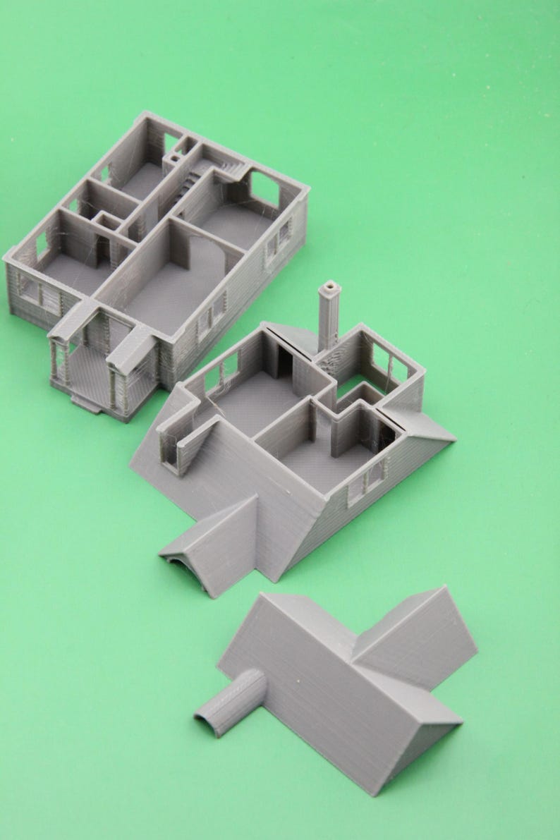 Op de afbeelding: Grijs 3D-geprint modelhuis in verschillende gedemonteerde secties, inclusief muren, dak en schoorsteen. Het model toont interieurdetails zoals kamers en trappen. De stukken zijn gerangschikt op een groene ondergrond.