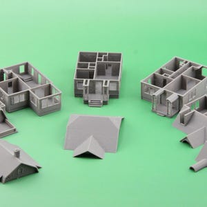 Op de afbeelding: Een verzameling grijze miniatuurhuismodellen, waaronder open structuren en losse dakonderdelen, is gerangschikt op een groene ondergrond. De modellen tonen interieurindelingen en architectonische details.