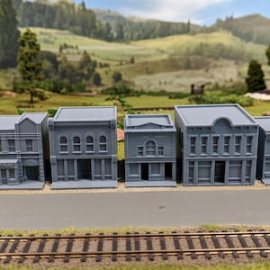 May include: A collection of five gray miniature buildings, including a Western Auto store, arranged on a gray surface with a railroad track in the foreground. The buildings have detailed architectural features.