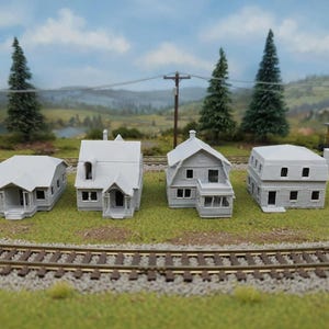 May include: Four miniature gray model houses of varying designs are arranged on a grassy surface with railroad tracks in the foreground. The houses include a bungalow, a two-story house, and a house with a porch. The background features trees and a yellow train car.