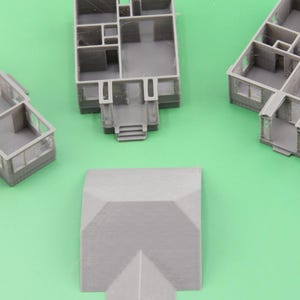 Op de afbeelding: Verschillende kleine, grijze, 3D-geprinte miniatuurhuismodellen en een dakstuk zijn gerangschikt op een groene ondergrond. De modellen bevatten gedetailleerde interieurindelingen, met muren, deuropeningen en ramen. Het dakstuk heeft een eenvoudig, schuin ontwerp.