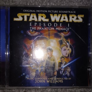 May include: A CD case for the Star Wars Episode I: The Phantom Menace soundtrack. The cover features a scene with characters and the text "STAR WARS EPISODE I THE PHANTOM MENACE". Music composed and conducted by John Williams.