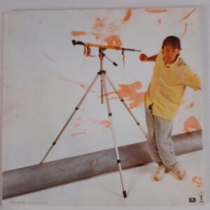 May include: A silver tripod with a yellow and black device attached, set against a white background with orange paint strokes. A person in a yellow shirt and gray pants is in the background.