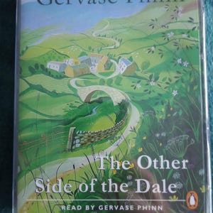 Op de afbeelding: Een doorzichtige plastic hoes met een luisterboek getiteld "The Other Side of the Dale" van Gervase Phinn. De omslag toont een groen landschap met een kronkelende weg, een stenen brug en gebouwen. Het Penguin-logo is zichtbaar.