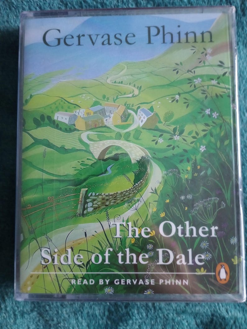 Op de afbeelding: Een doorzichtige plastic hoes met een luisterboek getiteld "The Other Side of the Dale" van Gervase Phinn. De omslag toont een groen landschap met een kronkelende weg, een stenen brug en gebouwen. Het Penguin-logo is zichtbaar.