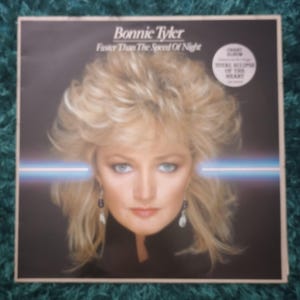 May include: Album cover for Bonnie Tyler's "Faster Than The Speed Of Night." The album art features a portrait of a woman with blonde hair and blue eyes. The title and artist's name are displayed at the top. A circular sticker on the right side advertises the album's chart success.
