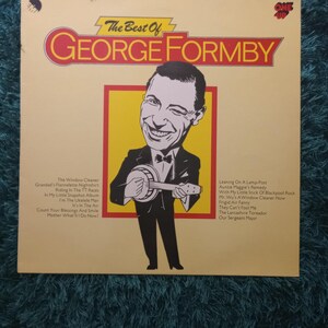 Puede incluir: Un álbum de vinilo vintage titulado "The Best Of George Formby". La portada del álbum presenta una ilustración de dibujos animados de George Formby tocando un banjo, sobre un fondo amarillo y rojo. Los títulos de las canciones están listados.