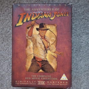 May include: The Adventures of Indiana Jones DVD movie collection box set. The cover features a brown and tan design with an image of Indiana Jones holding a whip. The text includes "Widescreen" and "Digitally Mastered for Superior Sound and Picture Quality."